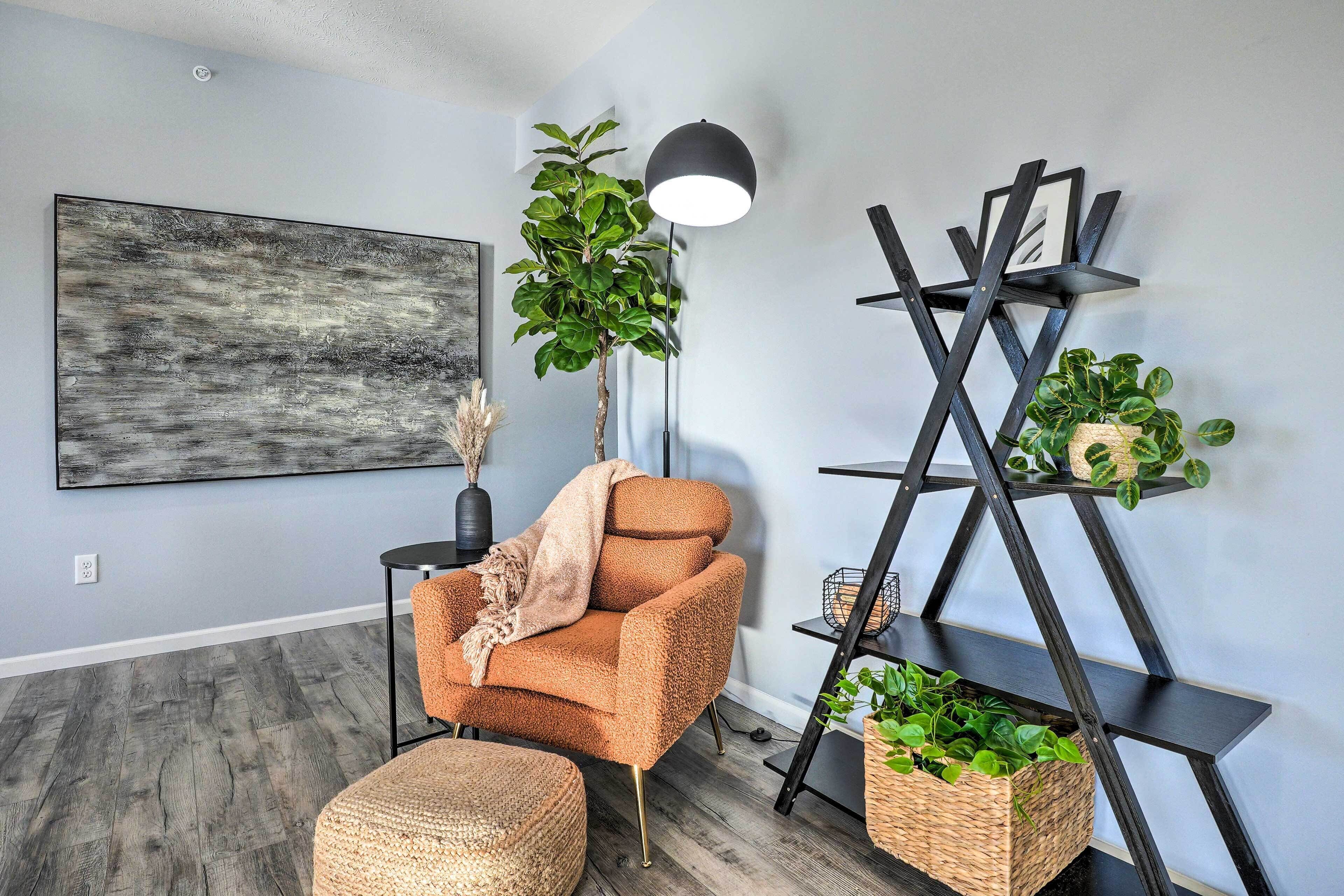 A modern living room with a grey wall, a large abstract painting, a brown armchair, a floor lamp, a black shelving unit with plants, and a woven basket on the floor.
