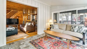 A spacious living room with a wooden staircase, a leather sofa, a beige couch, and a large window offering a view of the outdoors. - Center Ossipee Cabin w/ Game Room & Fire Pit! (Center Ossipee)