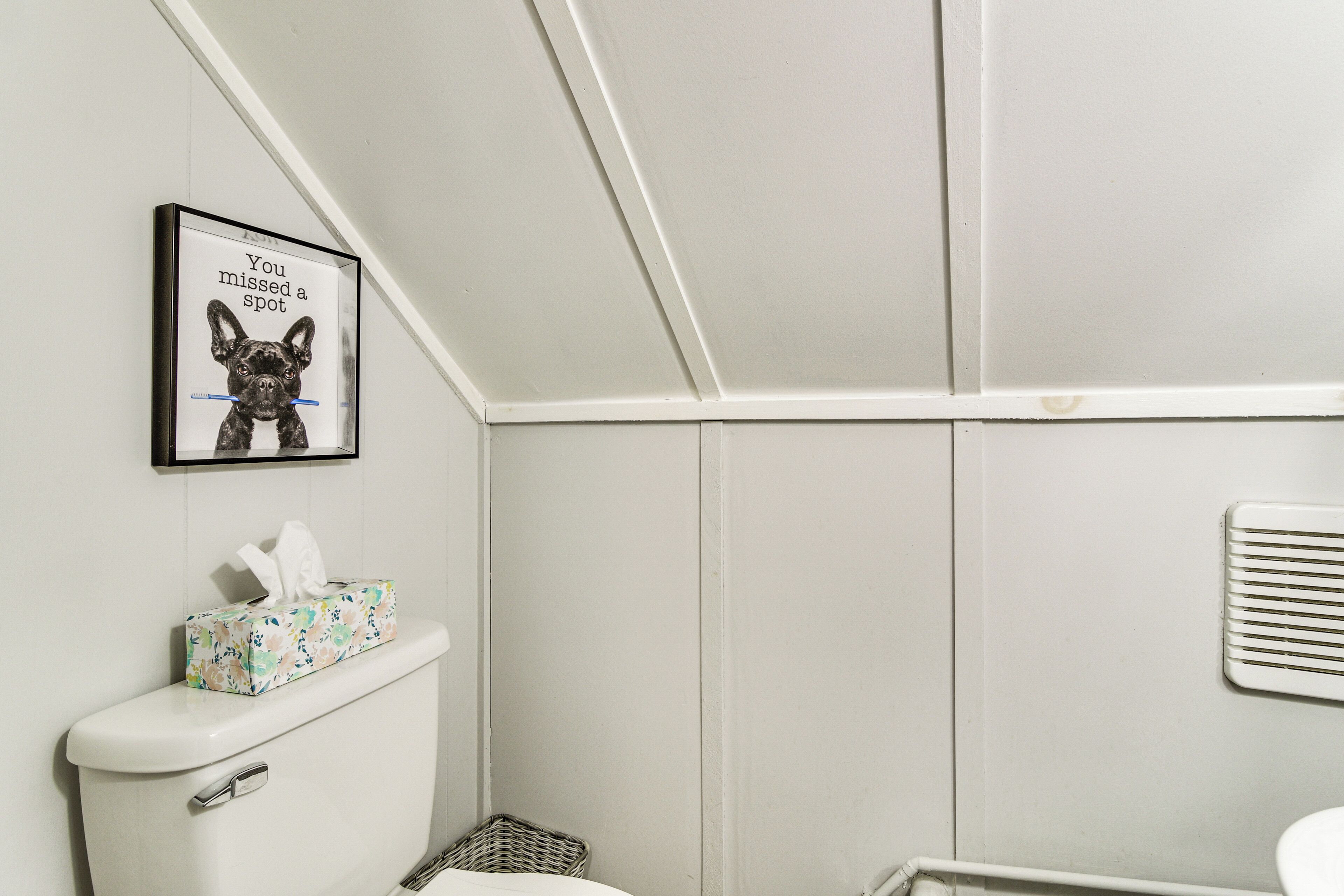 A bathroom with a toilet, a framed picture of a dog, a basket, and a vent.