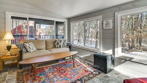 A cozy living room with a sofa, coffee table, and rug, large windows with a view of trees, and a lamp on a side table. - Center Ossipee Cabin w/ Game Room & Fire Pit! (Center Ossipee)