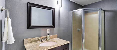 Un baño con lavamanos de encimera de granito, tocador de madera oscura, espejo y cabina de ducha de vidrio.