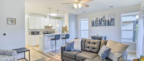 A modern living room with a grey sectional sofa, a patterned armchair, and a dark coffee table. A kitchen with white cabinets and a marble backsplash is visible in the background.