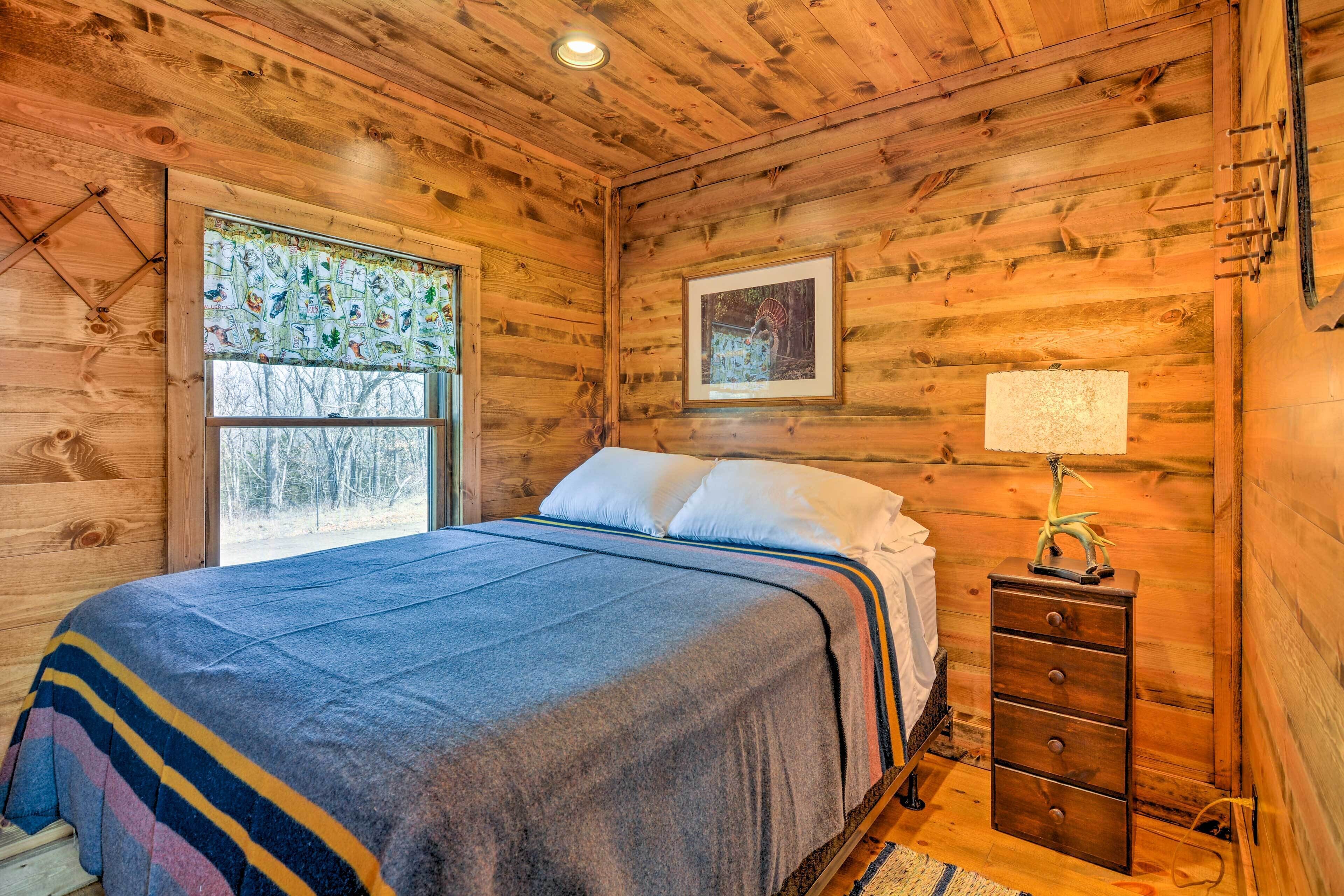 A wooden cabin bedroom with a bed, a dresser, a window with a view of trees, and a framed picture on the wall.