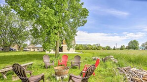 Un espace extérieur aménagé avec des chaises Adirondack et un foyer, entouré d’un champ herbeux et d’arbres.