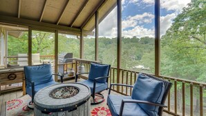 A covered outdoor seating area with a fire pit table, two blue cushioned chairs, and a view of a forested landscape. - Walk to Lake: Osage Beach Home w/ Grill! (Osage Beach)