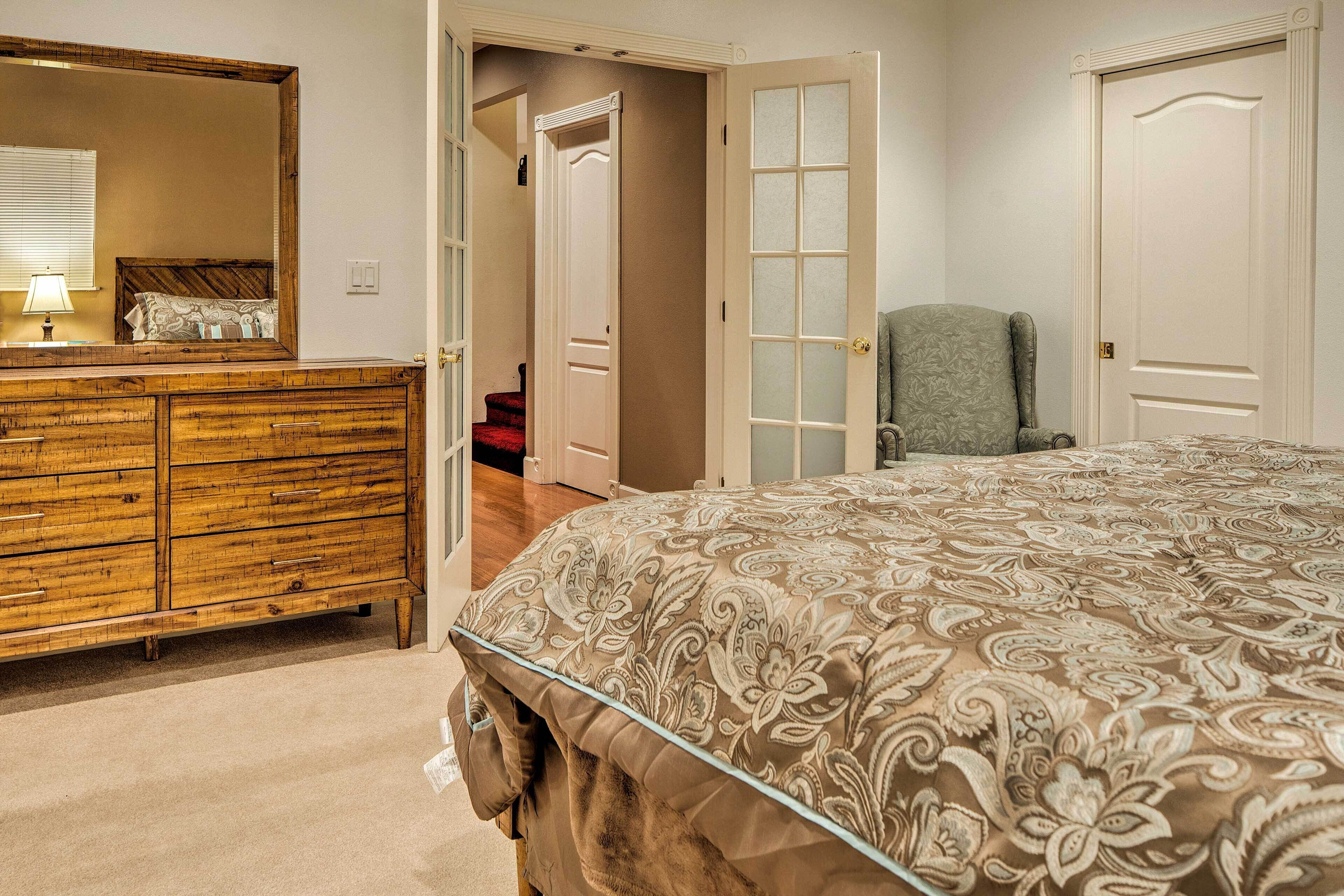 A bedroom with a large bed, a wooden dresser, a chair, and a door leading to another room.
