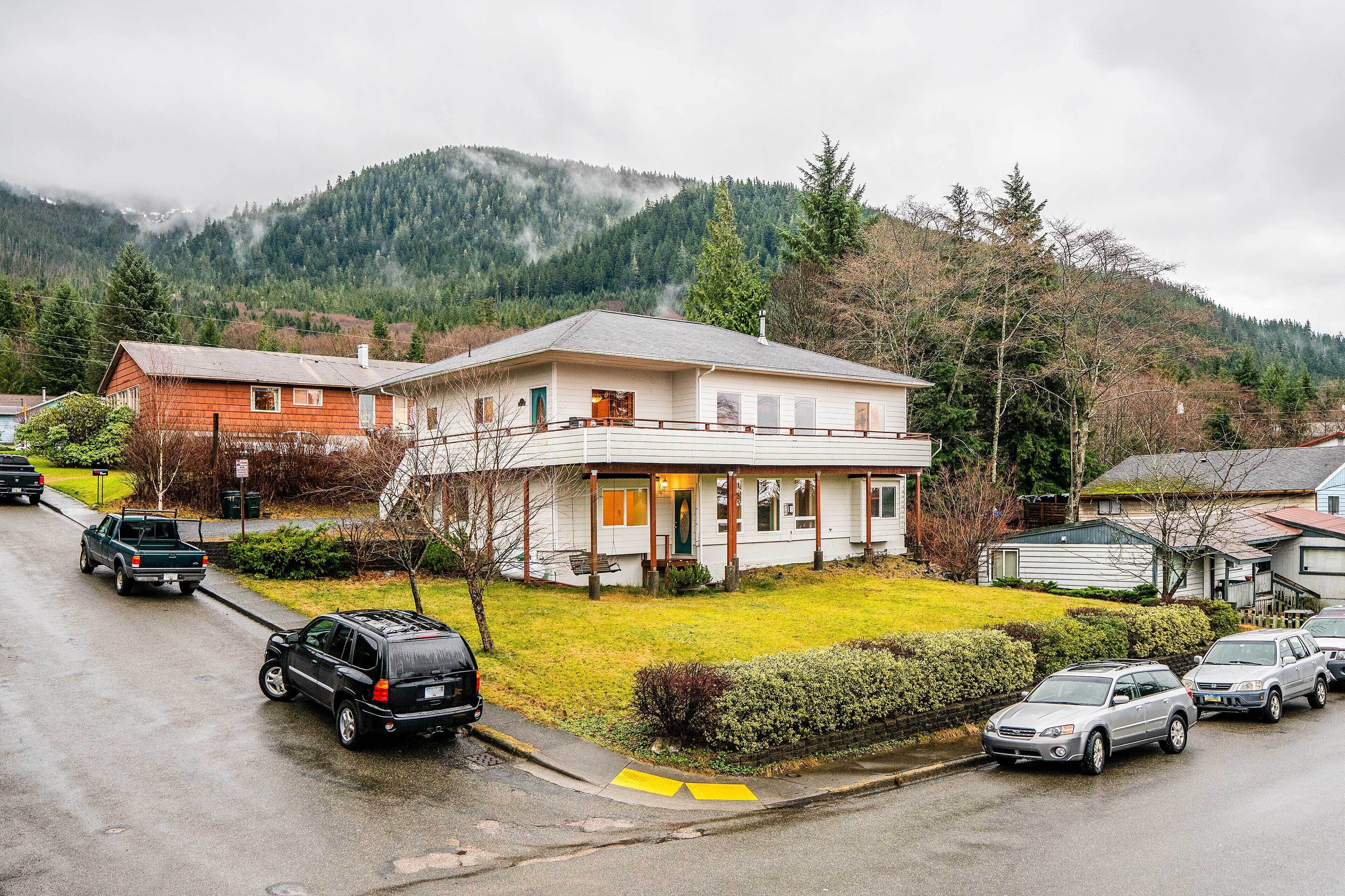 A two-story house with a front porch, parked cars, and a mountain backdrop.