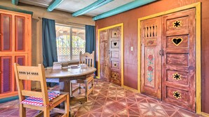 Una habitación con una mesa de comedor de madera, sillas y un suelo estampado. Hay una puerta con detalles decorativos y una ventana con cortinas.
