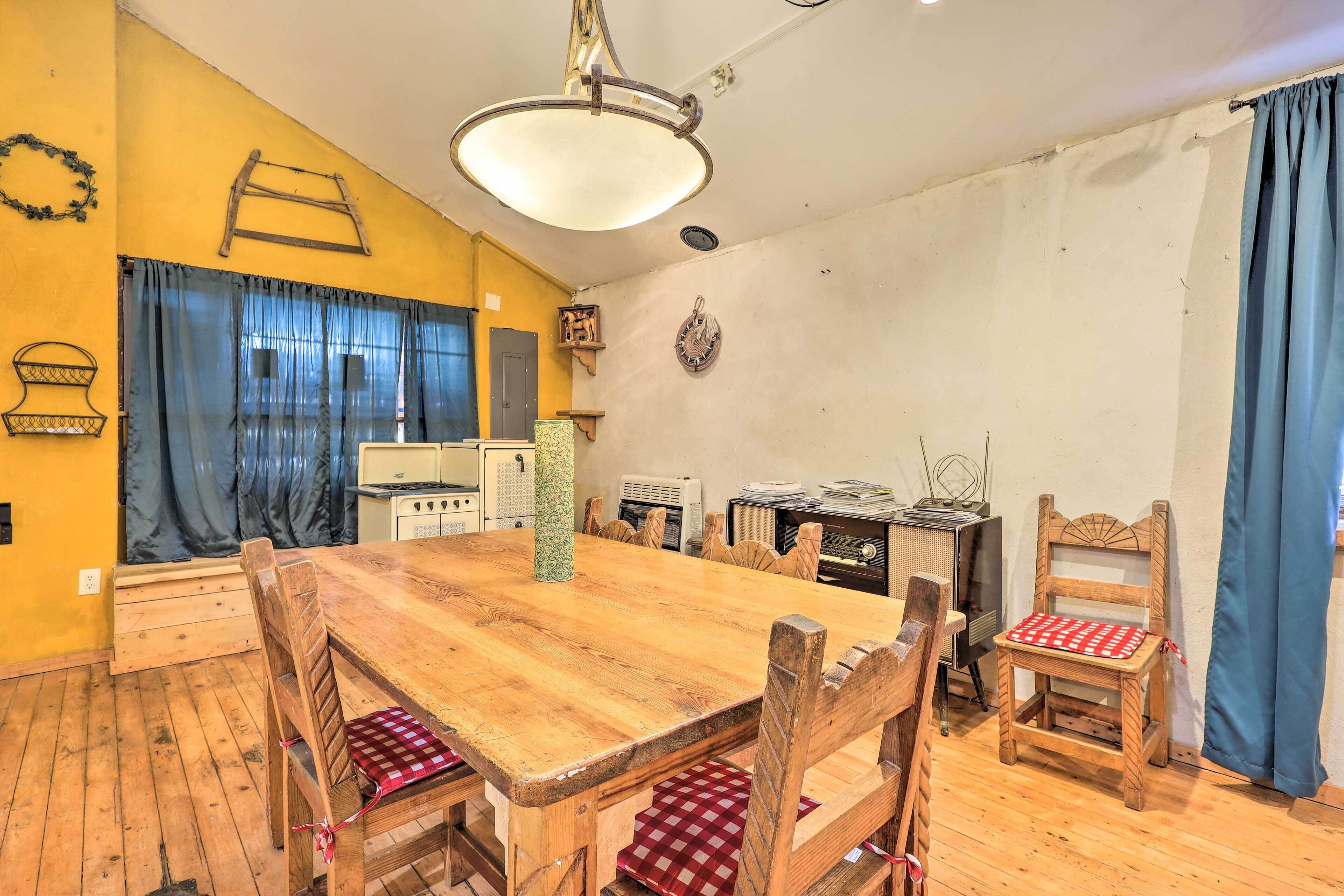 A dining area with a wooden table and chairs, a hanging light fixture, and a kitchen in the background.