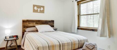 Un dormitorio con cabecera de madera, cama con ropa de cama a rayas, mesita de noche con lámpara y ventana con cortinas.