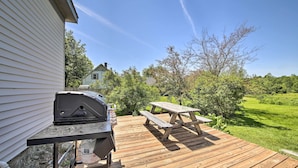 A wooden deck with a grill and picnic table, surrounded by greenery and a house in the background. - Near Lakes, Bay & Trails! Historic Waldoboro Gem (Waldoboro)