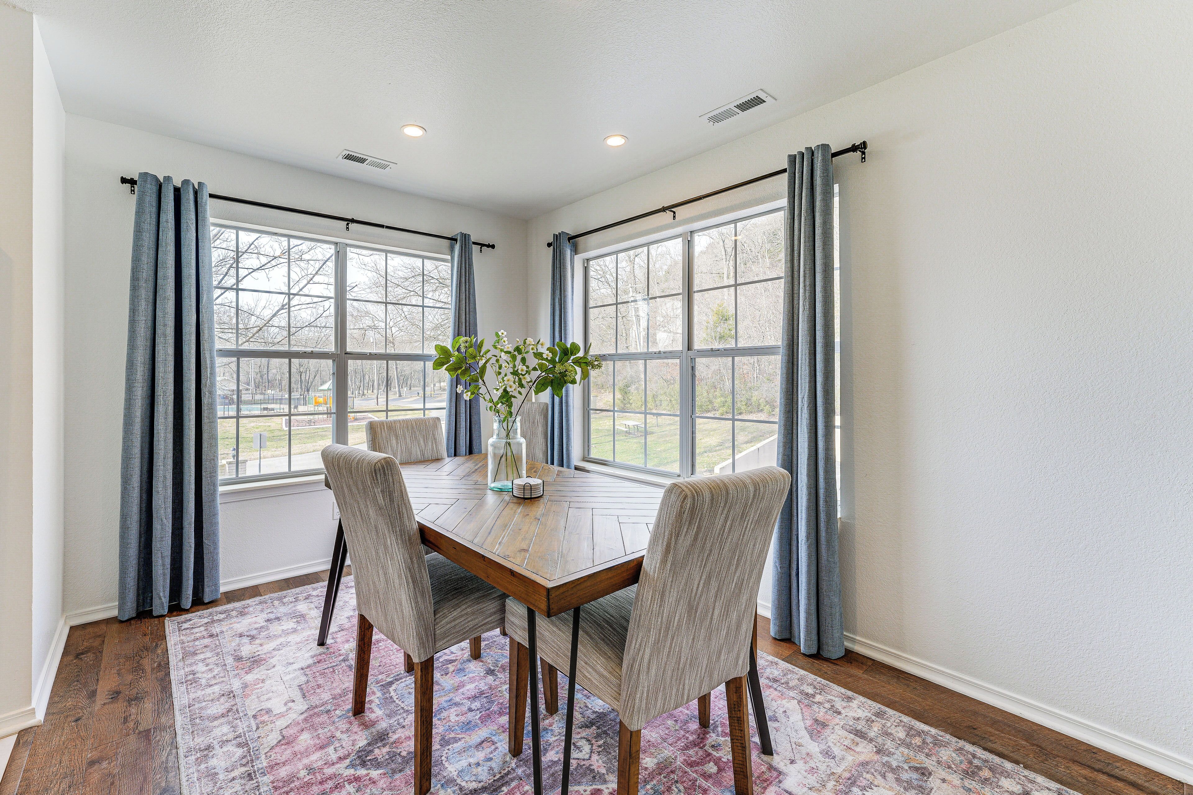 A dining area with a wooden table, four chairs, a vase with flowers, and large windows with curtains.