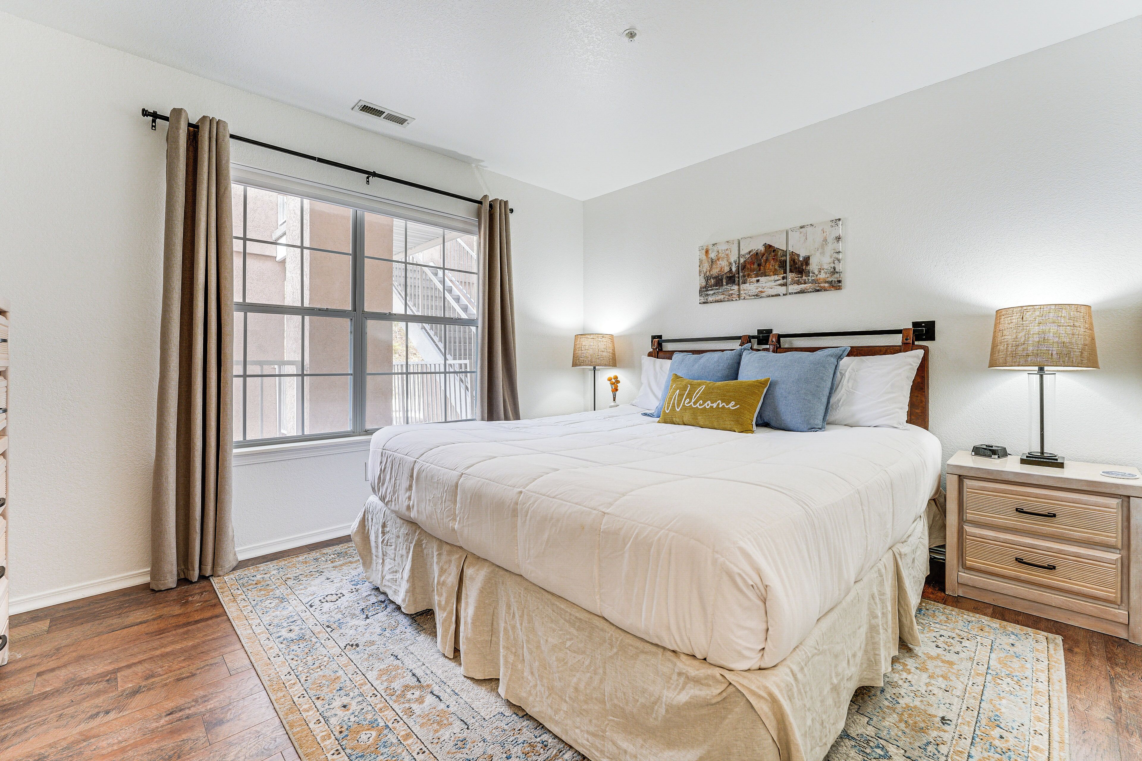 A bedroom with a large bed, a nightstand with a lamp, a window with curtains, and a wall-mounted artwork.