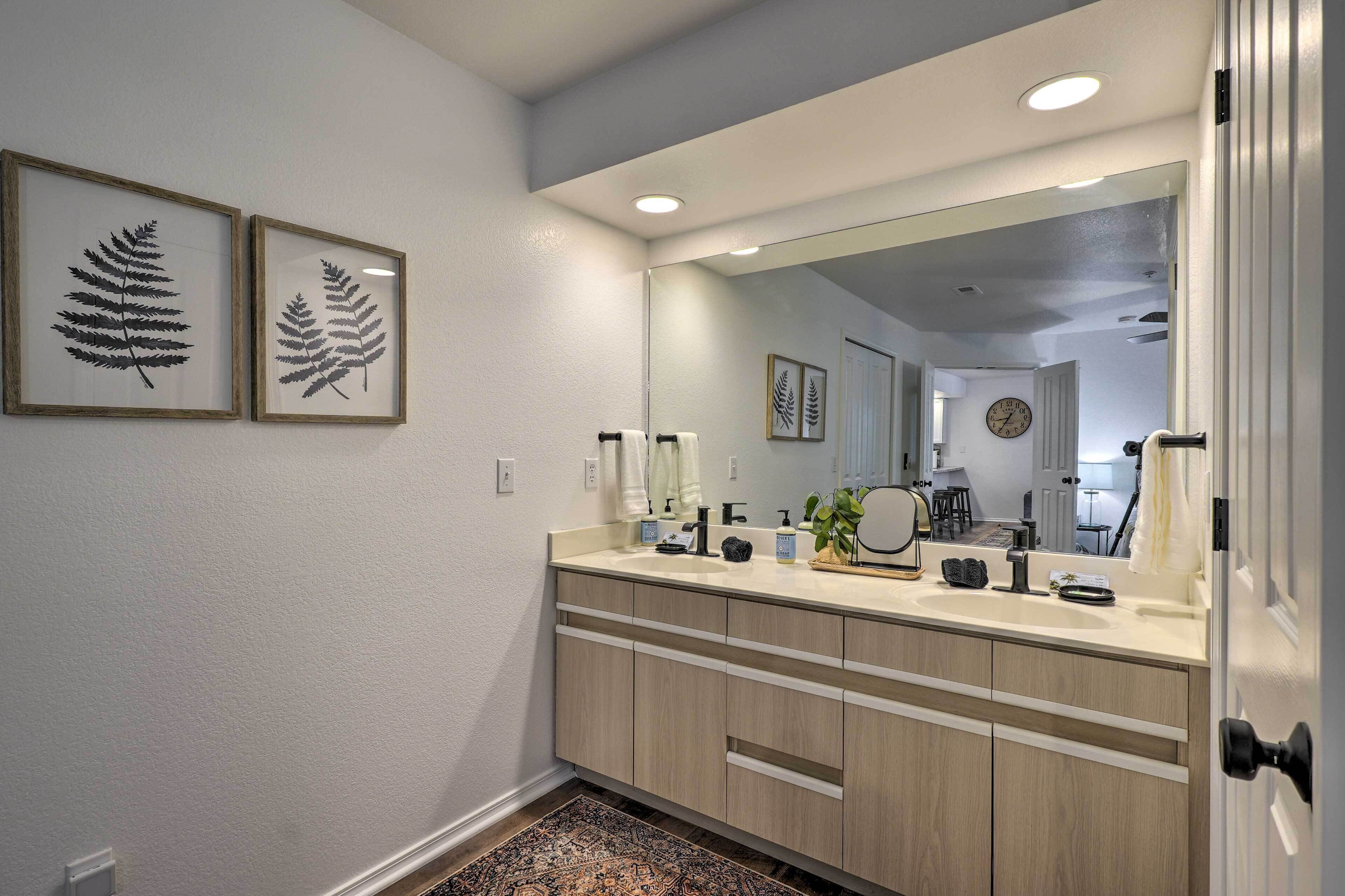 A modern bathroom with a large mirror, two framed fern pictures on the wall, and a beige countertop with various toiletries.