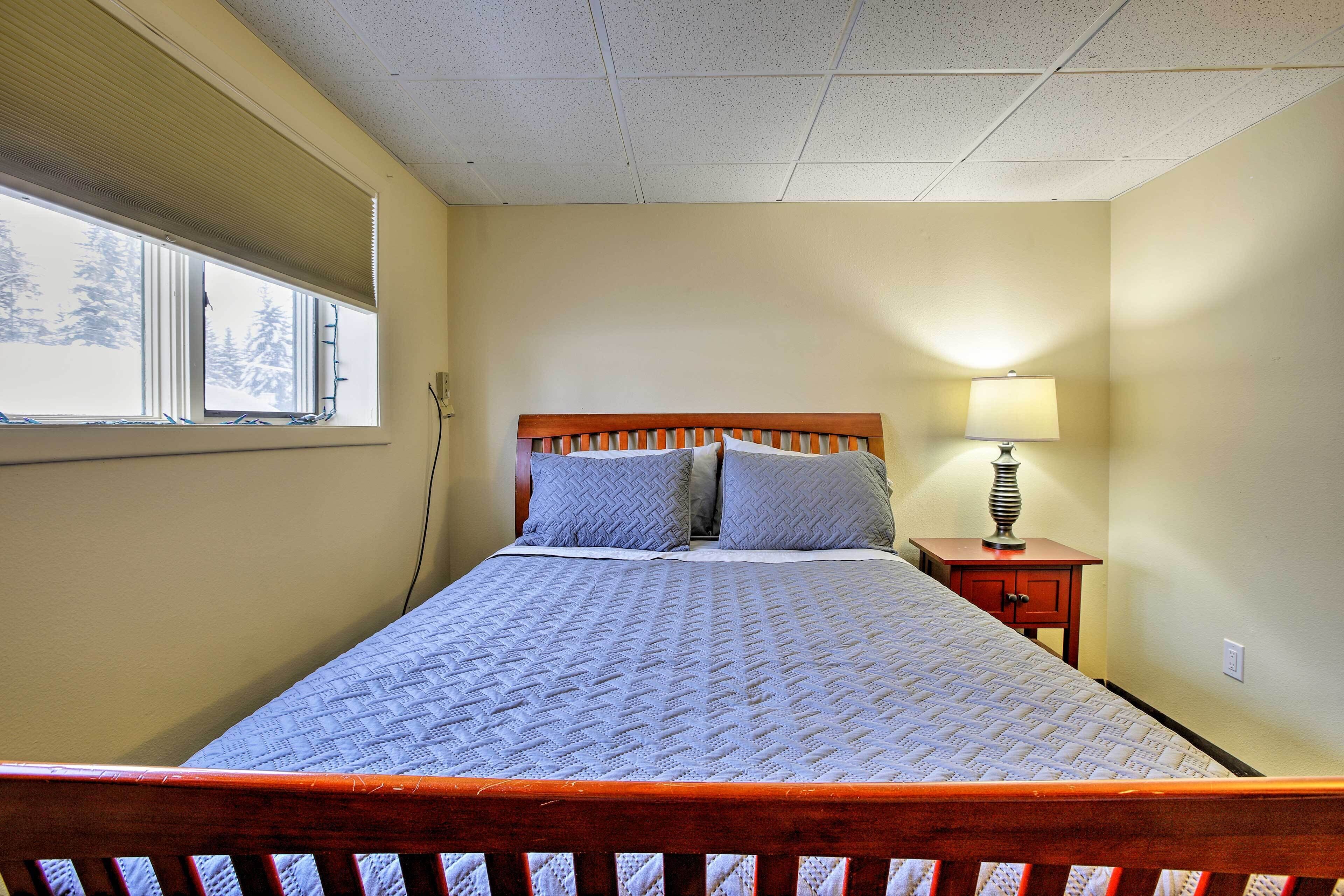 A bedroom with a bed, a nightstand with a lamp, a window with blinds, and a wall socket.