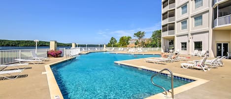 A swimming pool with clear blue water, surrounded by lounge chairs and a building with balconies.