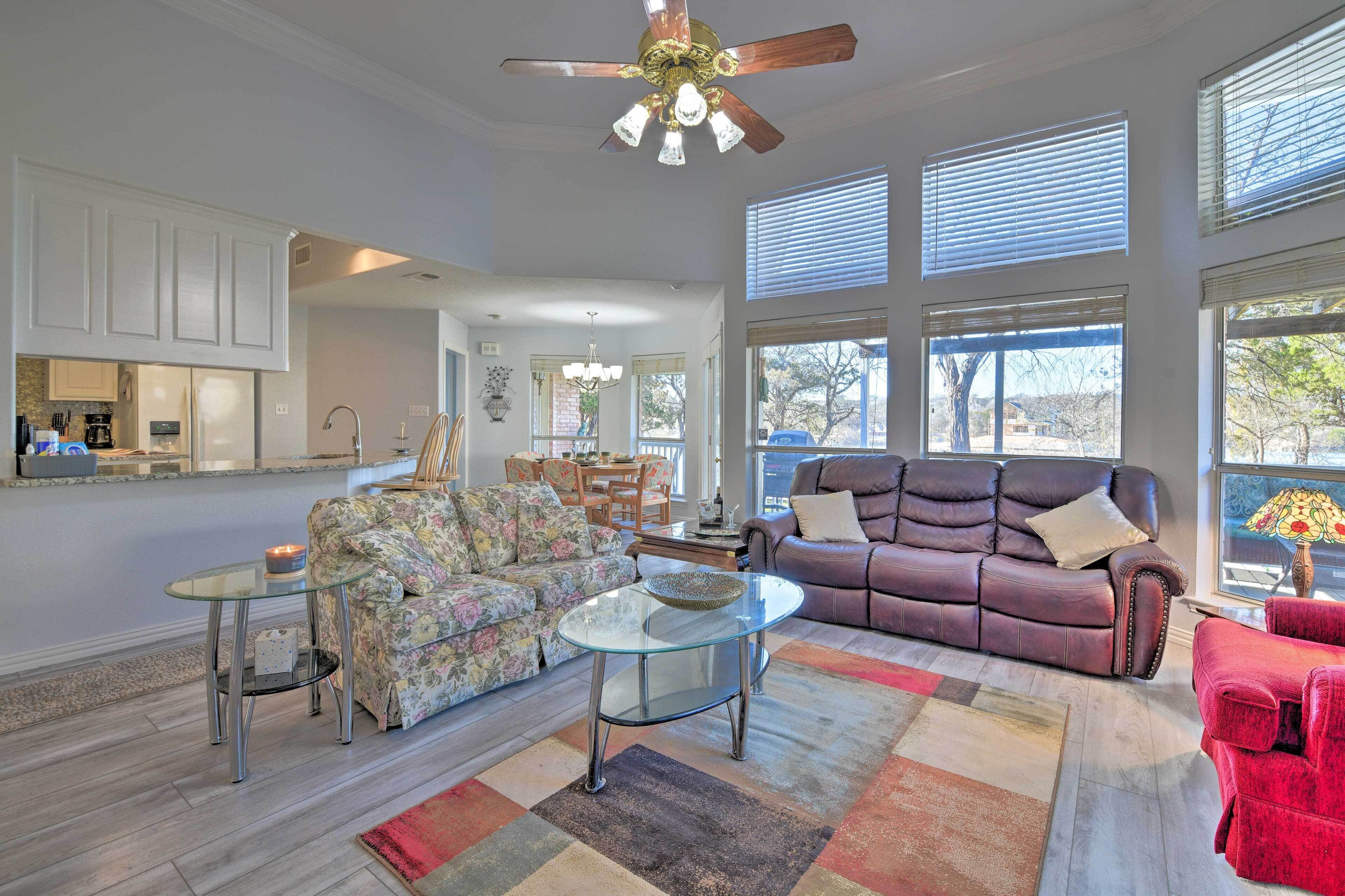 A spacious living room with a glass coffee table, a leather sofa, and a red armchair. A kitchen is visible in the background with white cabinets and a refrigerator.