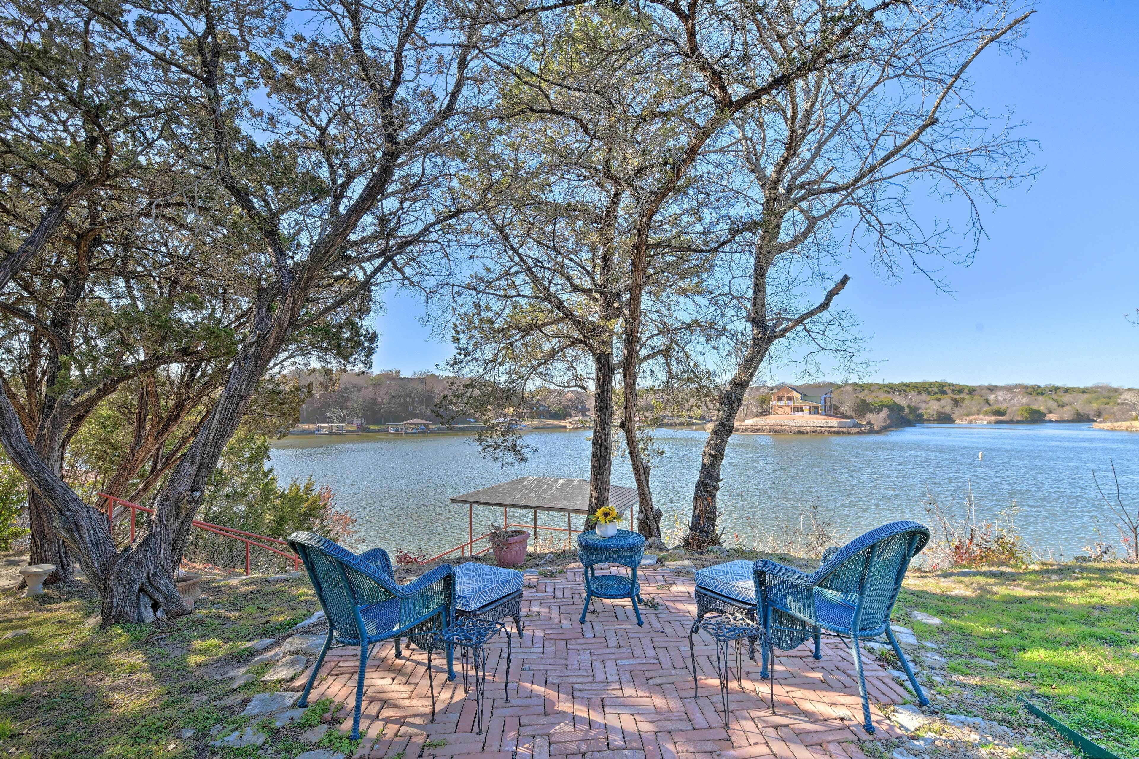A patio with wicker chairs and a table overlooking a lake, with trees and a distant house visible.