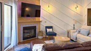 A living room with a fireplace, a television mounted above it, a blue armchair, a beige sofa, and a coffee table.
