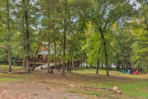 A cabin with a deck and stairs leading to a porch, surrounded by trees and a grassy area with Adirondack chairs.
