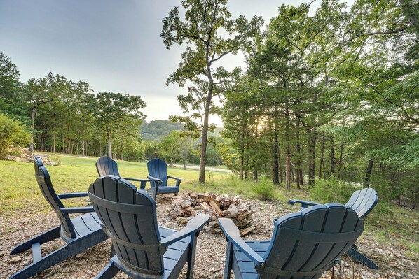 A fire pit with four Adirondack chairs arranged around it, set in a wooded area with a grassy field in the background.