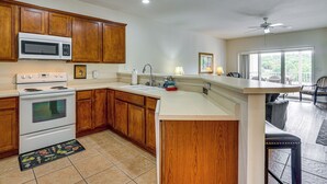 A kitchen with wooden cabinets, a microwave, stove, sink, and a bar area with a chair. - Lakefront Osage Beach Condo: Dock Your Boat Here! (Osage Beach)