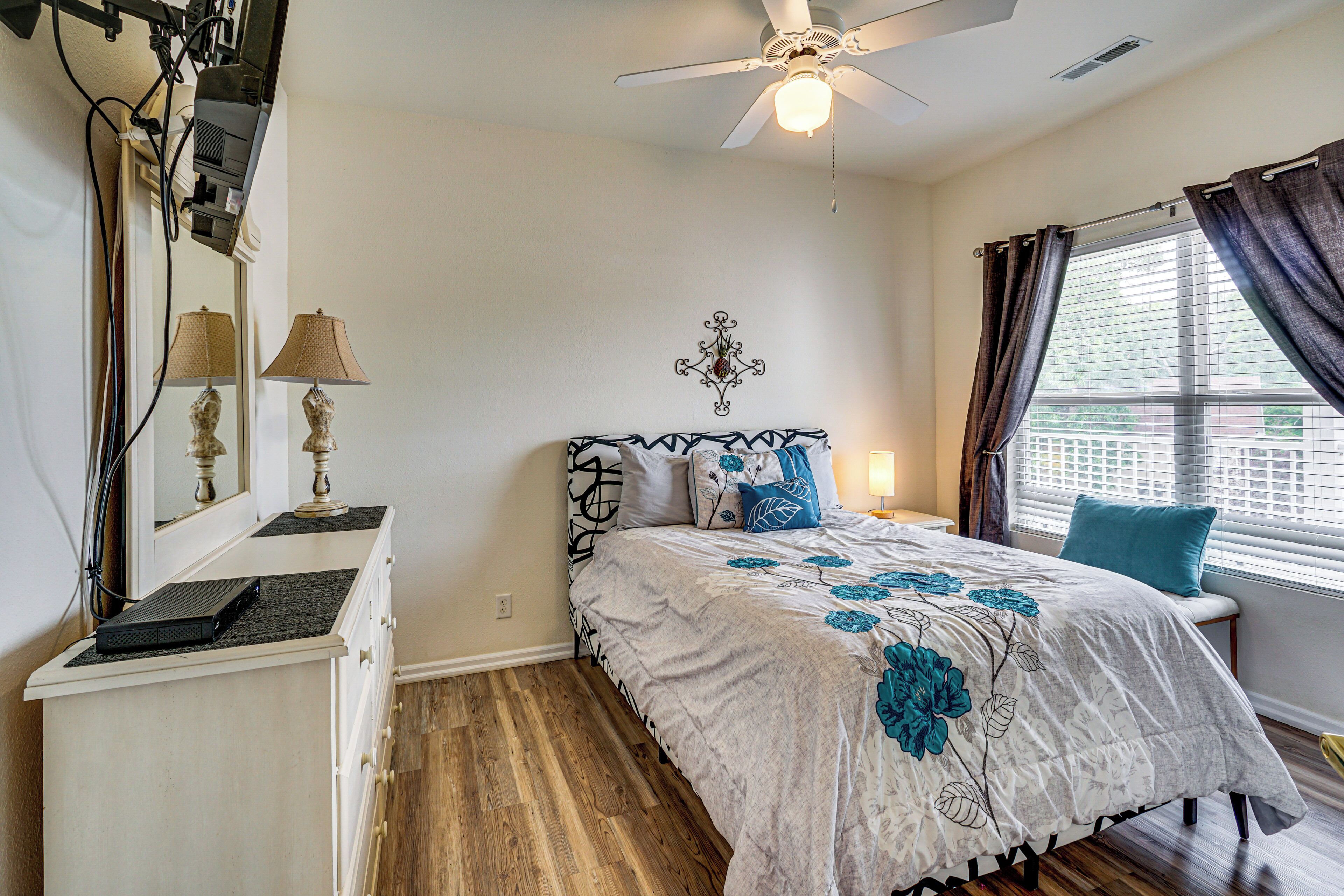 A bedroom with a bed, bedside table, lamp, ceiling fan, and a window with curtains.