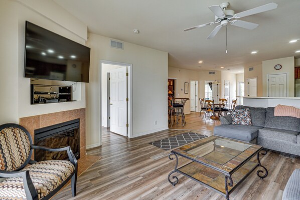A spacious living room with a fireplace, a flat-screen TV, a ceiling fan, and a dining area in the background. - Lakefront Osage Beach Condo: Dock Your Boat Here! (Osage Beach)