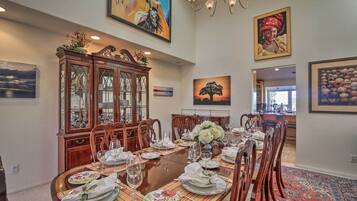 A dining room with a large wooden table set for six, surrounded by chairs. There is a wooden cabinet with glass doors to the left, and a chandelier above the table.