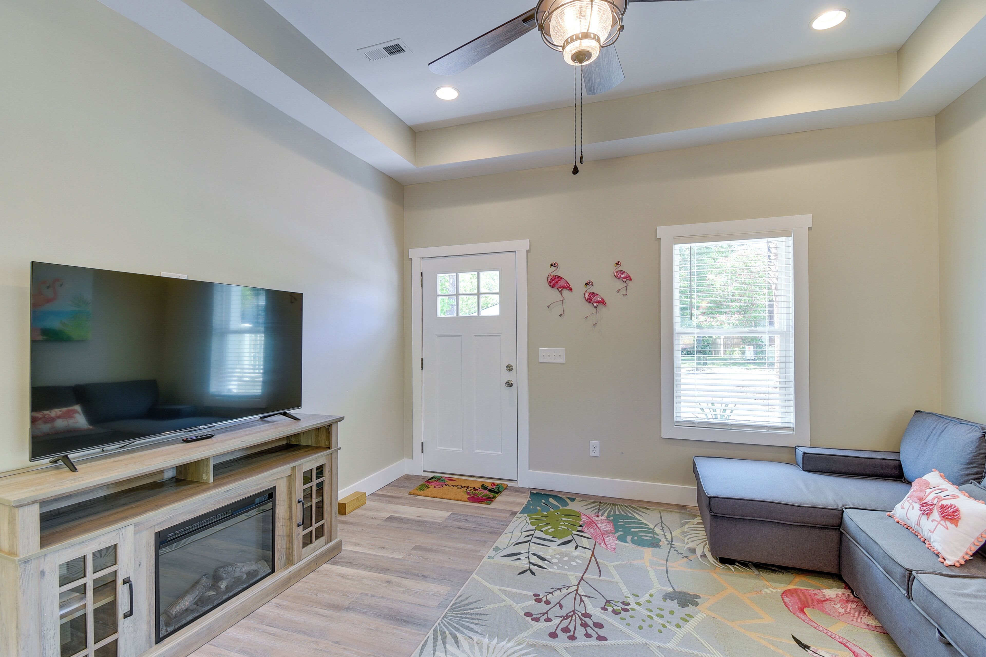 A living room with a flat-screen TV on a wooden stand, a ceiling fan, a door with a window, and a sofa with a cushion.