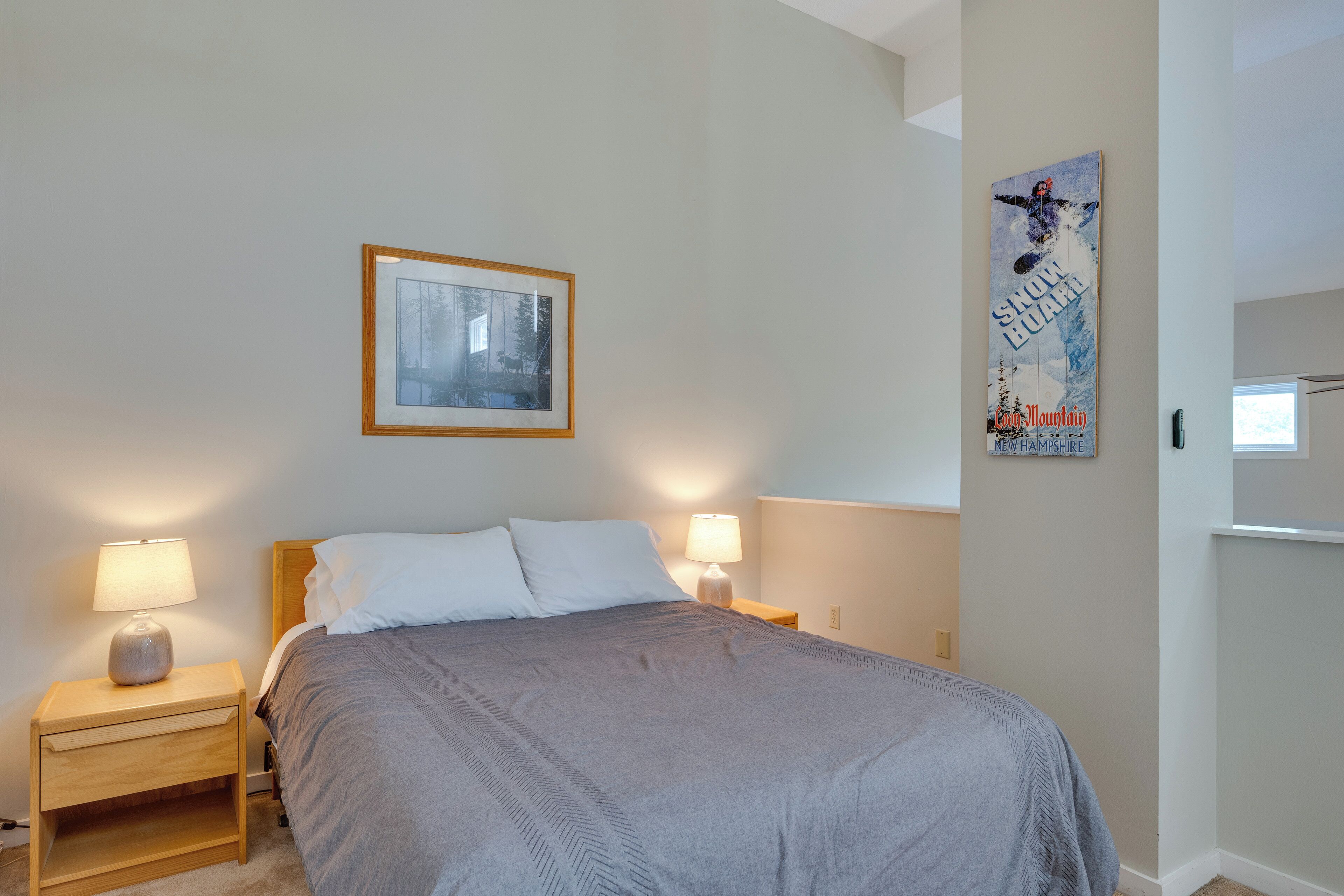 A neatly made bed with a grey blanket, two bedside lamps, a framed picture on the wall, and a poster advertising a ski resort.