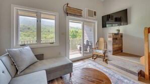 Un salón con sofá, una mesa de centro de madera, televisor montado en la pared y vistas al exterior a través de un ventanal corredero.