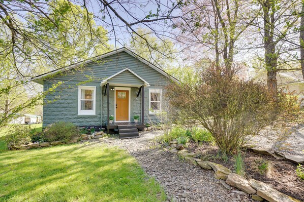A small, single-story house with a front porch, a wooden door, and a small garden area with plants and a stone pathway.