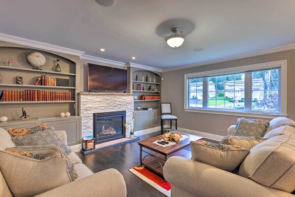 A living room with a fireplace, bookshelves, a sofa, and a window with a view. - 25 Mi to Nyc: Walkable Long Island Apt w/ Balcony (Glen Cove)