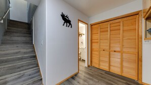 Un couloir avec un escalier en bois, une décoration murale représentant un cerf et une armoire en bois dotée de portes à persiennes.