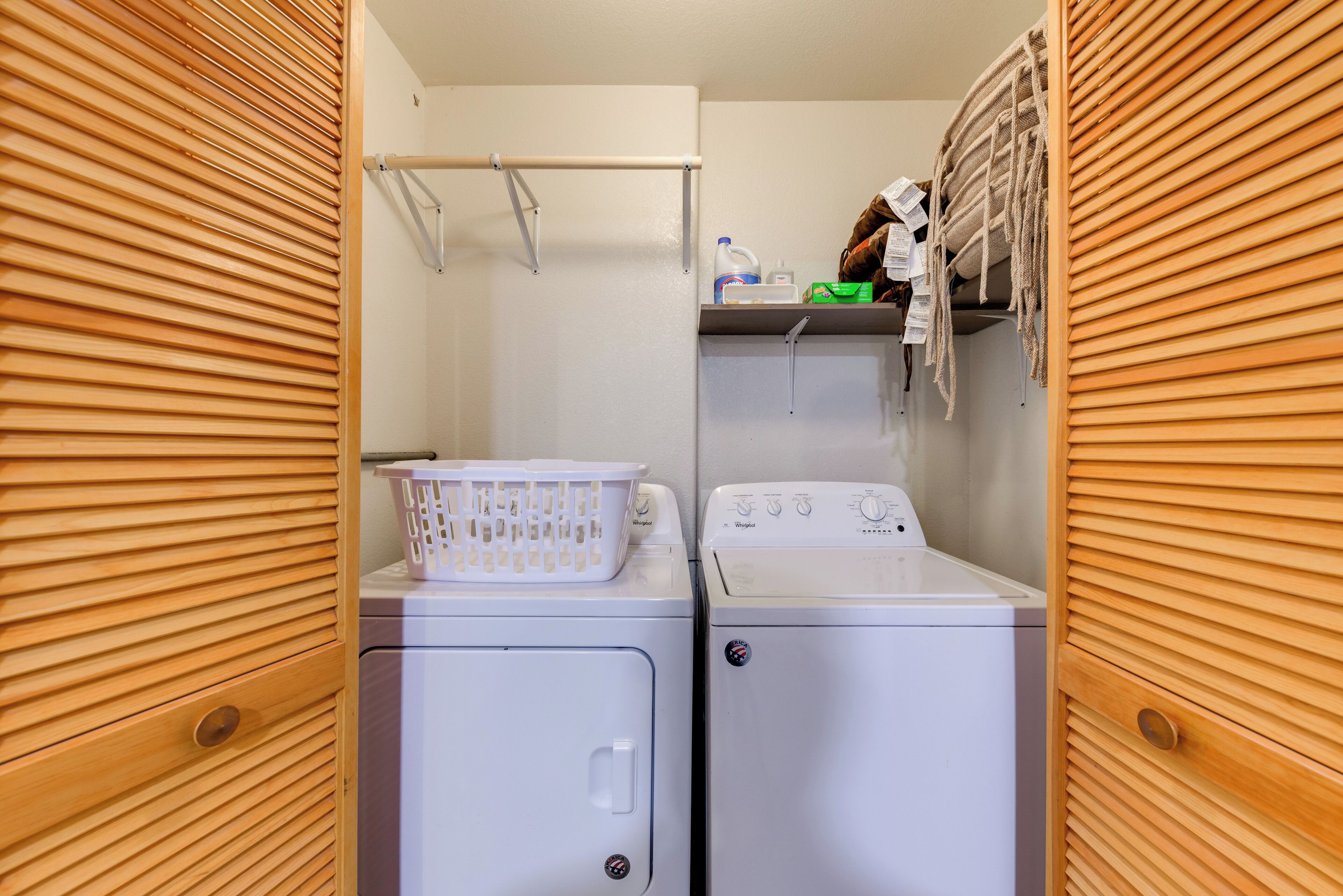 A laundry room with two front-loading washers, a drying rack, and a shelf with cleaning supplies.