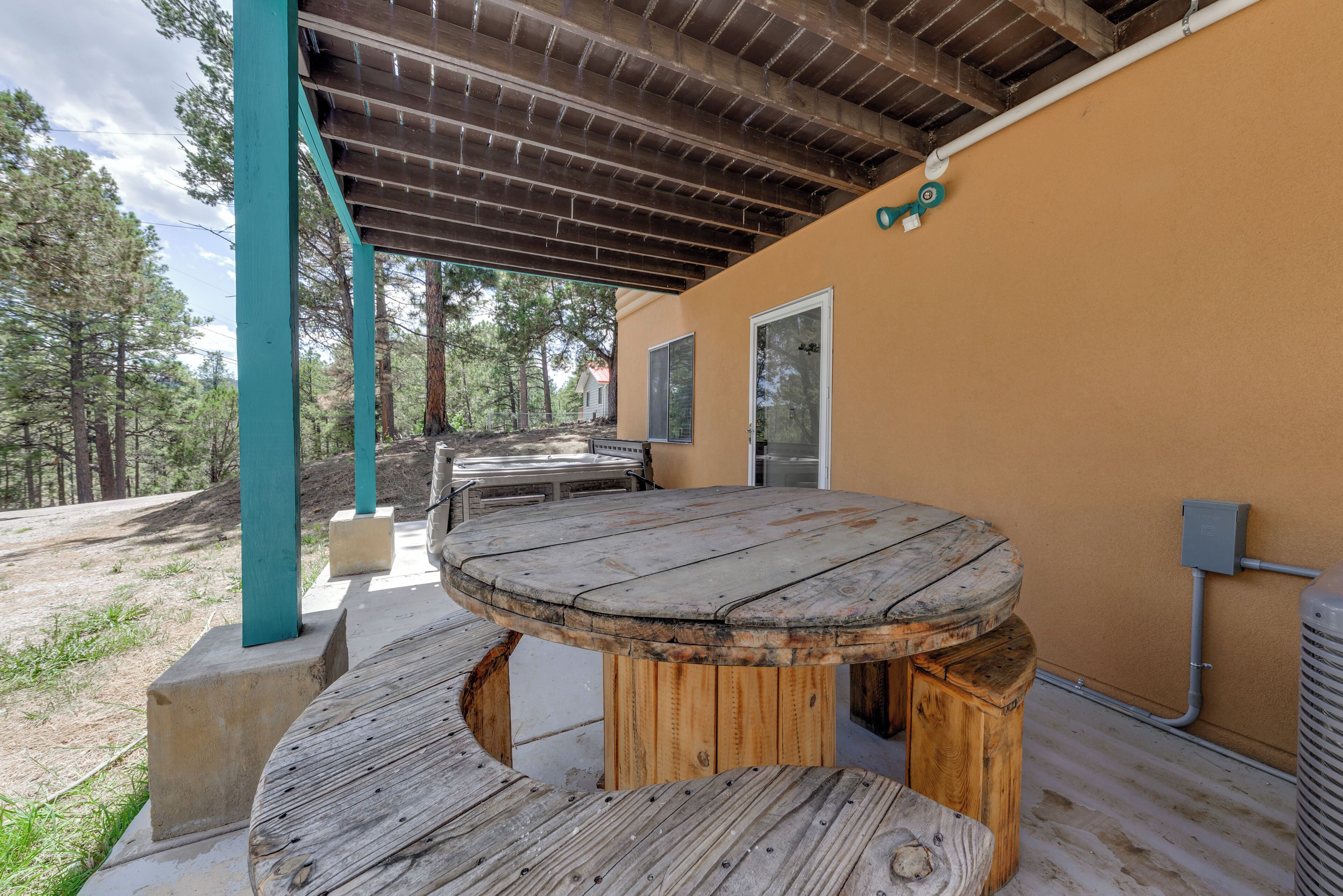 A wooden outdoor table and bench under a covered patio area with a view of trees and a building in the background.