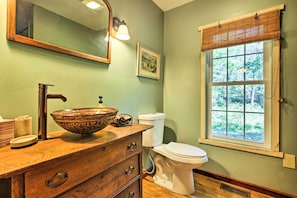 Une salle de bain avec un meuble-lavabo en bois, un grand lavabo en forme de cuve, des toilettes, une fenêtre donnant sur la verdure et un tableau encadré au mur.