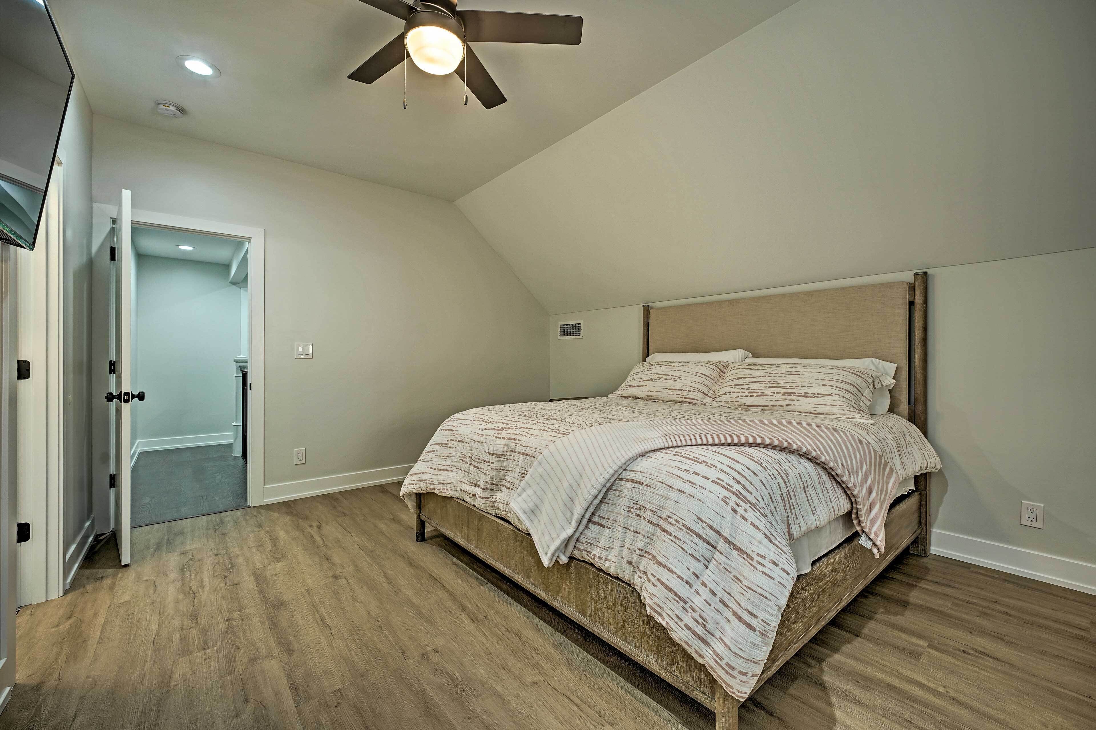 A bedroom with a bed, wooden flooring, a ceiling fan, and a door leading to another room.