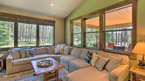 A cozy living room with a sectional sofa, a wooden coffee table, and large windows offering a view of a forested area. - Modern-rustic Ronald Getaway: Hot Tub, Grill! (Cle Elum)
