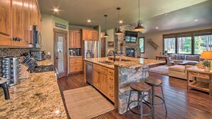 A spacious kitchen with granite countertops, wooden cabinets, and a stone island. There is a stone fireplace, a large window, and a cozy seating area with a sofa and bar stools. - Modern-rustic Ronald Getaway: Hot Tub, Grill! (Cle Elum)
