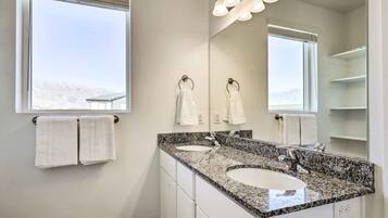A bathroom with two sinks, a large mirror, and a window with towels hanging on the wall.