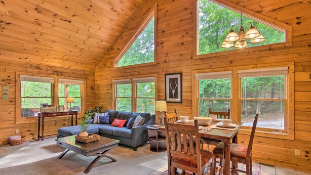 A cozy cabin interior with a sofa, dining table, and chairs, large windows, and wooden walls. - Hot Tub Soaks! 'moonshine Manor' Near Lake Lure (Lake Lure)