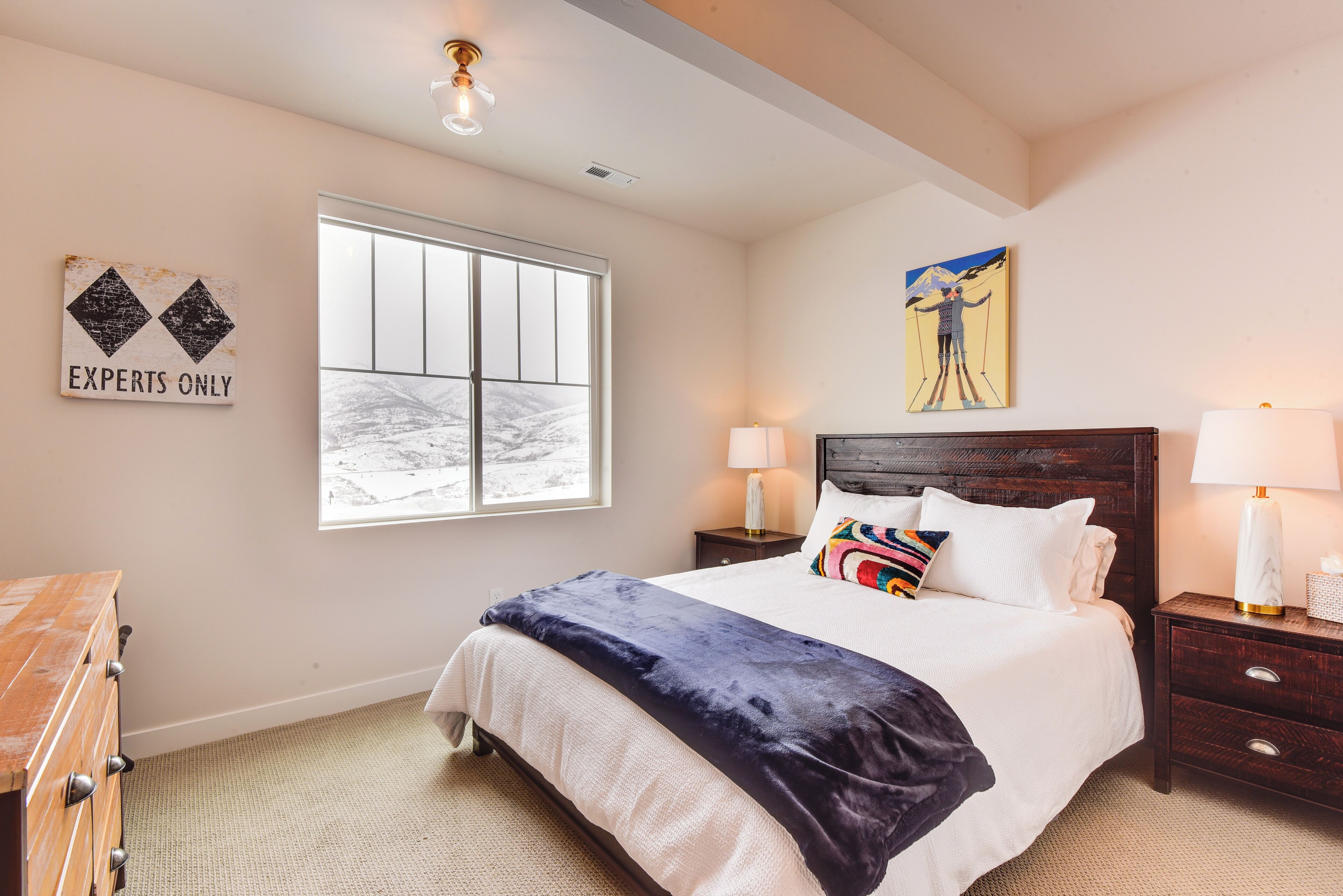 A bedroom with a large bed, a wooden dresser, a window with a view, a painting, and a wall sign.