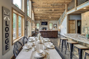 A spacious lodge interior with a dining area set for a meal, a stone fireplace, and a kitchen with a large island.