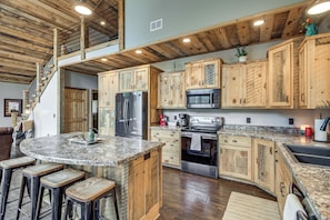 A kitchen with wooden cabinets, stainless steel appliances, a granite countertop, and a bar area with stools. - Modern Lakeside Chalet on Crow Wing Chain w/ Dock! (Nevis)