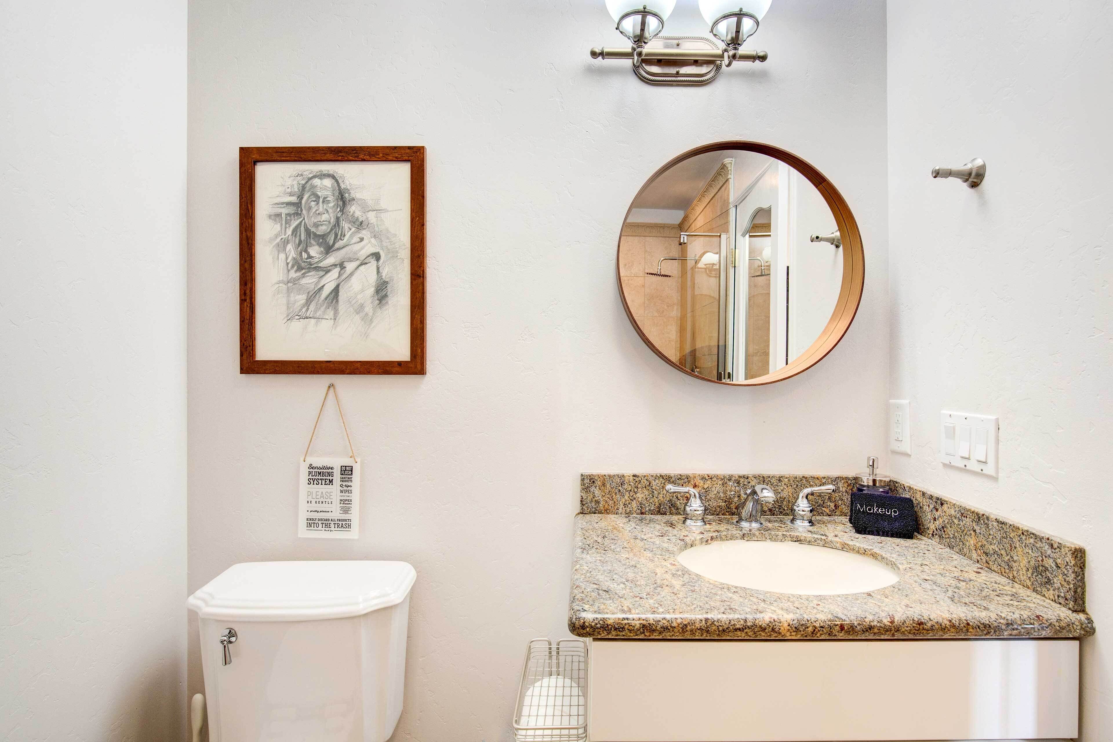 A bathroom with a white toilet, a granite sink, a framed picture, a round mirror, and a hanging light fixture.