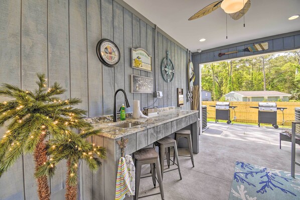 A kitchen with a wooden wall, bar stools, a ceiling fan, and a view of a backyard with a grill.