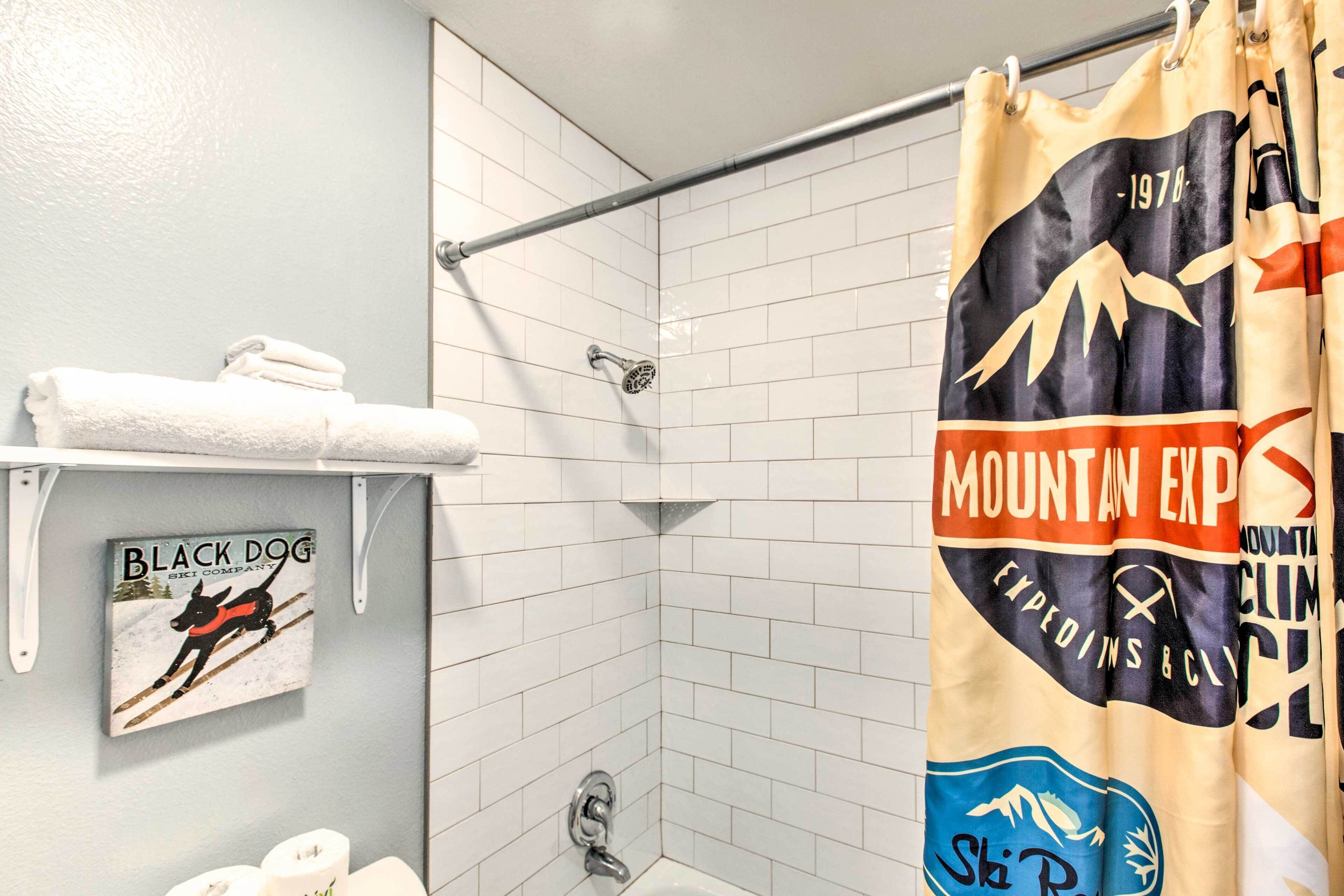 Un espace douche avec un mur recouvert de carreaux blancs, une tringle de douche en métal et un rideau de douche orné d’un motif de montagne.