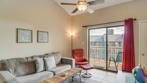 A living room with a sofa, a red chair, a wooden coffee table, and a colorful ottoman. There is a ceiling fan, a floor lamp, and a view of a balcony with a railing.